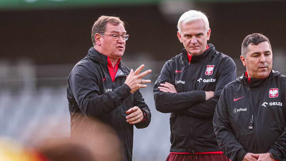 Trzech mężczyzn w czarnych kurtkach sportowych z białymi elementami i logo Polski, prawdopodobnie sztab szkoleniowy, stoi na tle zielonego boiska.