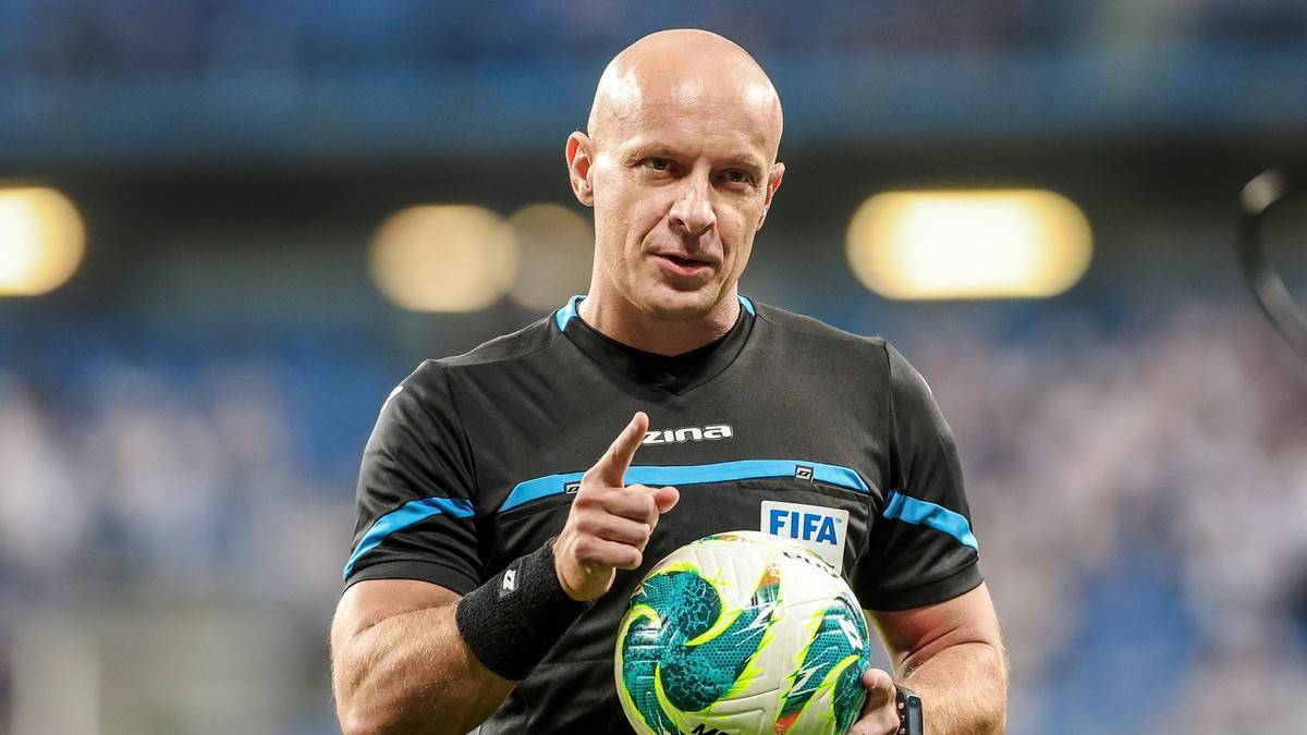Szymon Marciniak, polski sędzia piłkarski, ubrany w czarną koszulkę sędziowską z niebieskimi wstawkami i logo FIFA, trzymający w ręku piłkę nożną.