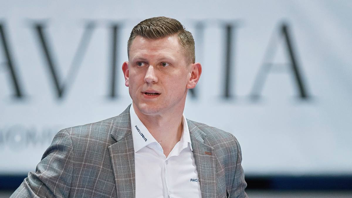 Mężczyzna w białej koszuli i marynarce w kratkę, z krótkimi blond włosami, patrzy w bok z lekkim uniesieniem brwi. W tle rozmazany napis.