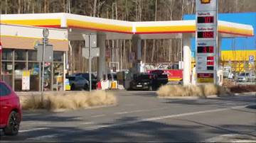 Obraz przedstawia stację paliw Shell z widocznymi dystrybutorami i zaparkowanymi samochodami. Widoczny jest słup z cenami paliw.