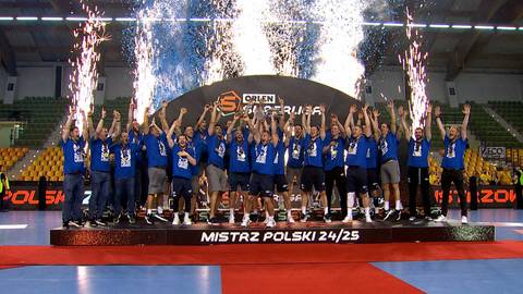 Drużyna piłkarzy ręcznych w niebieskich strojach stoi na podium, świętując zwycięstwo. W tle widać tablicę z napisem "MISTRZ POLSKI 24/25", a nad głowami zawodnik&oacute;w unoszą się fontanny fajerwerk&oacute;w. Na trybunach siedzą kibice.