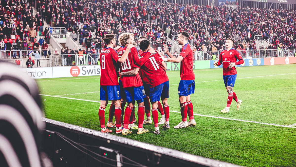 Piłkarze w czerwonych strojach świętują gola na boisku piłkarskim, na trybunach zgromadzeni kibice.