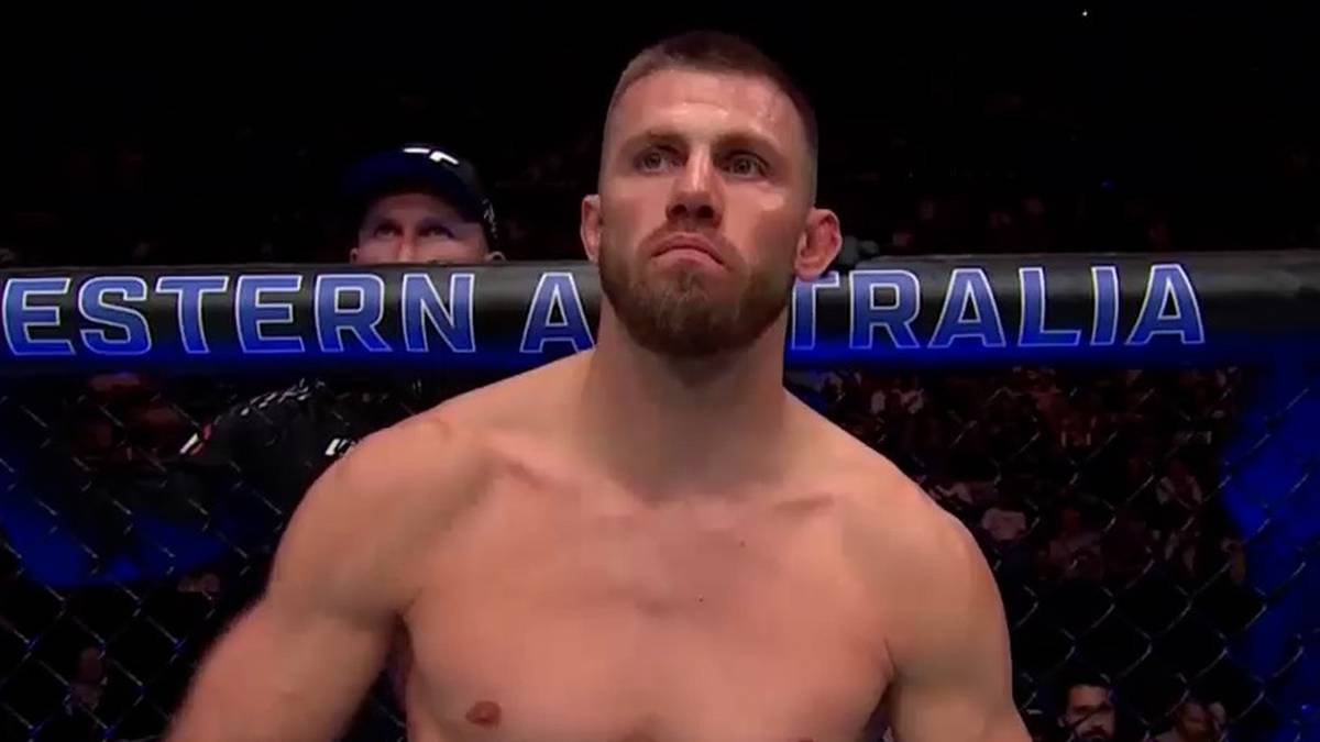 Zawodnik MMA z kr&oacute;tką brodą, patrzący przed siebie. W tle widoczna reklama "WESTERN AUSTRALIA" i zarysy publiczności.