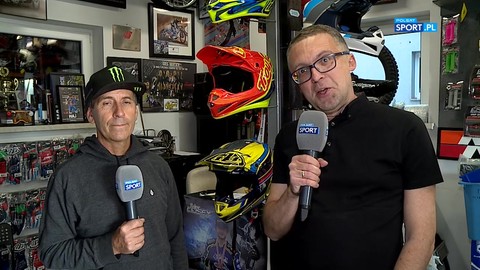 Dwóch mężczyzn w pomieszczeniu pełnym kasków motocyklowych i pucharów. Mężczyzna po lewej ma na głowie czapkę z daszkiem, a mężczyzna po prawej nosi okulary. Obaj trzymają mikrofony.