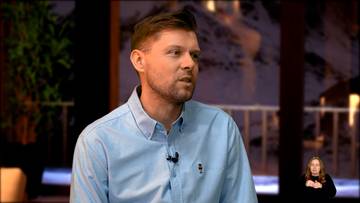 Mężczyzna w jasnoniebieskiej koszuli z kołnierzykiem, m&oacute;wiący do kamery. Na jego piersi znajduje się mikrofon. W tle widać fragment rośliny oraz oświetlone okno z widokiem na krajobraz g&oacute;rski.