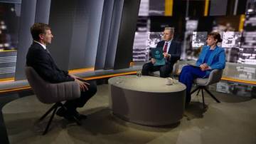 Trzy osoby siedzące przy okrągłym stoliku w studiu telewizyjnym. Mężczyzna po lewej w garniturze rozmawia z dwiema osobami po prawej. Kobieta po prawej ma na sobie niebieską marynarkę.