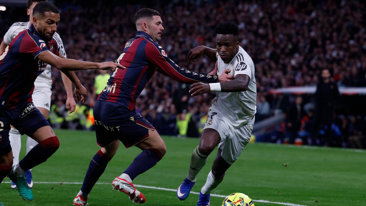 Piłkarz w białej koszulce prowadzi piłkę, ścigany przez dw&oacute;ch piłkarzy w czerwono-niebieskich strojach.