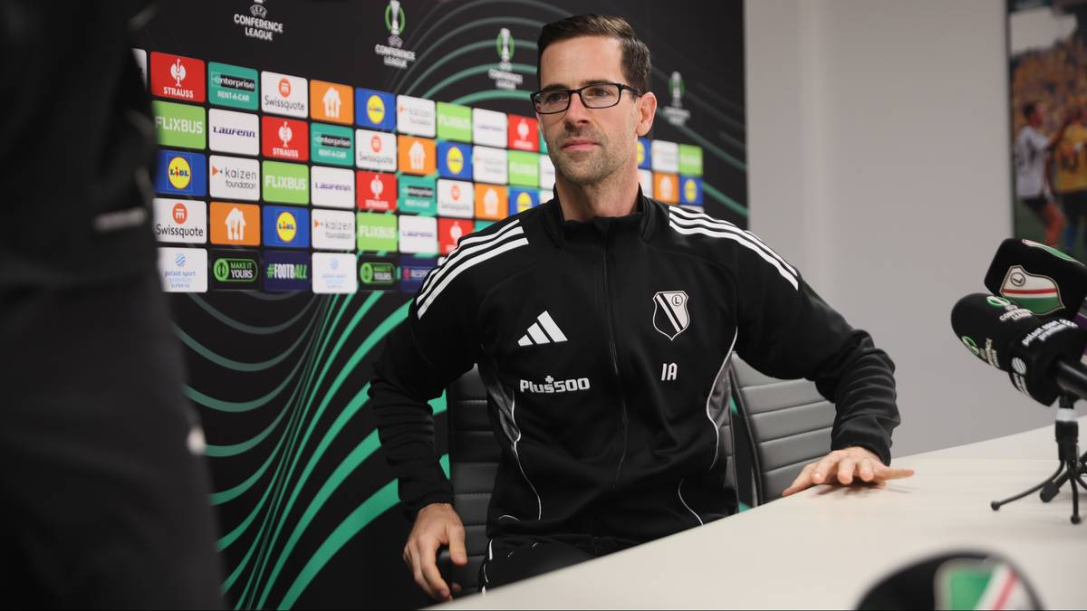 Trener Legii Warszawa siedzi przy stole podczas konferencji prasowej, ubrany w dres z logo klubu.