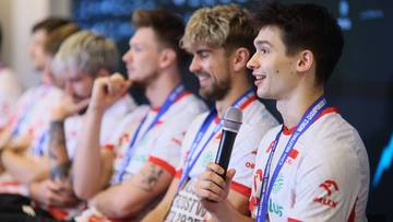 Grupa młodych mężczyzn siedzących w rzędzie, niekt&oacute;rzy z nich noszą białe koszulki z niebieskimi akcentami i medale na szyjach. Jeden z mężczyzn trzyma mikrofon.