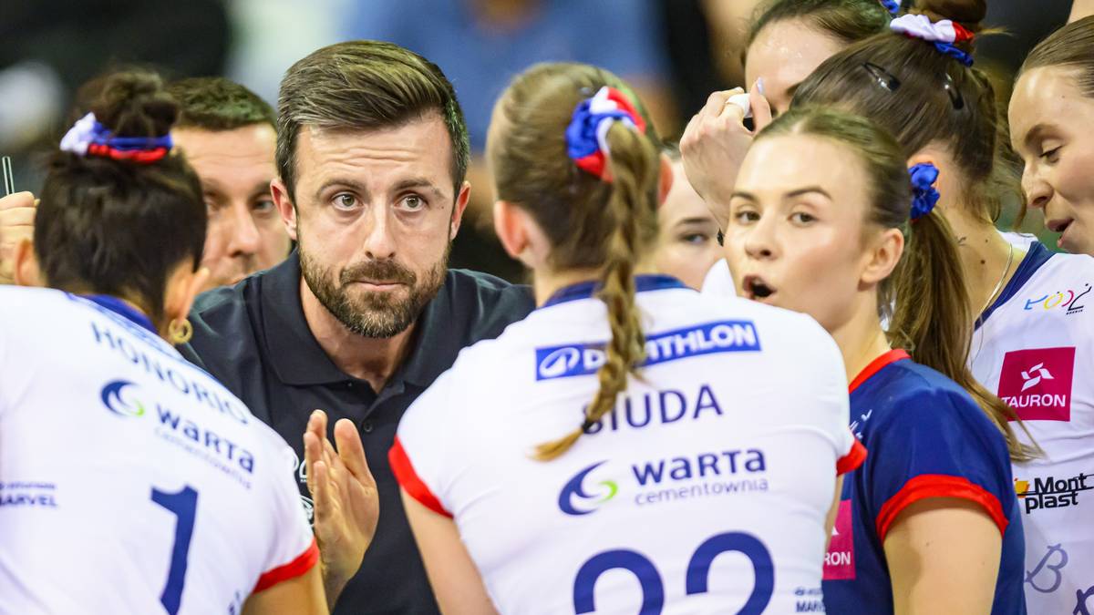 Trener siatkówki w czarnej koszulce, otoczony przez zawodniczki w białych i niebieskich strojach sportowych, przekazuje instrukcje podczas przerwy w meczu.