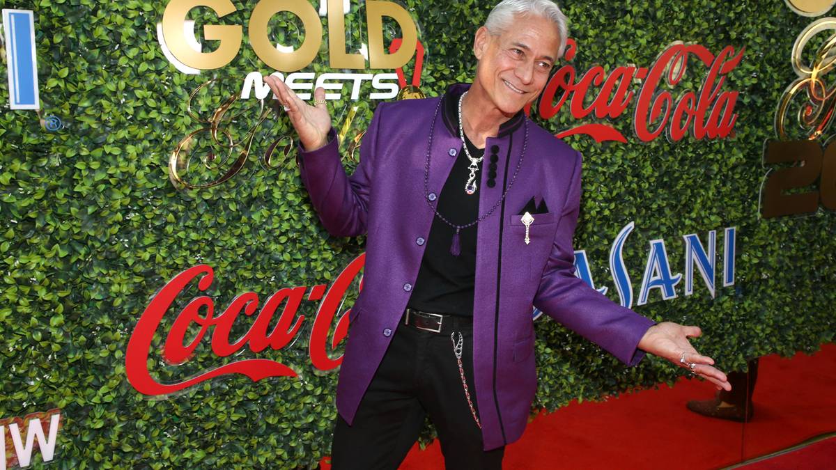 Mężczyzna w fioletowej marynarce stoi przed ścianą z bluszczu z widocznymi logotypami "GOLD MEETS" i "Coca-Cola". Gestykuluje rękami w geście powitania lub prezentacji.