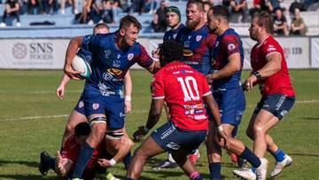Grupa zawodników rugby w niebieskich i czerwonych strojach walcząca o piłkę na trawiastym boisku.