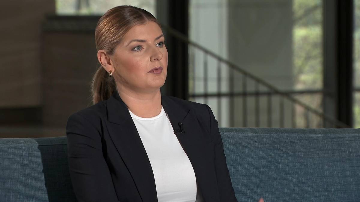 Kobieta w ciemnej marynarce i białej koszuli m&oacute;wi, siedząc na kanapie. W tle widać schody i okna.