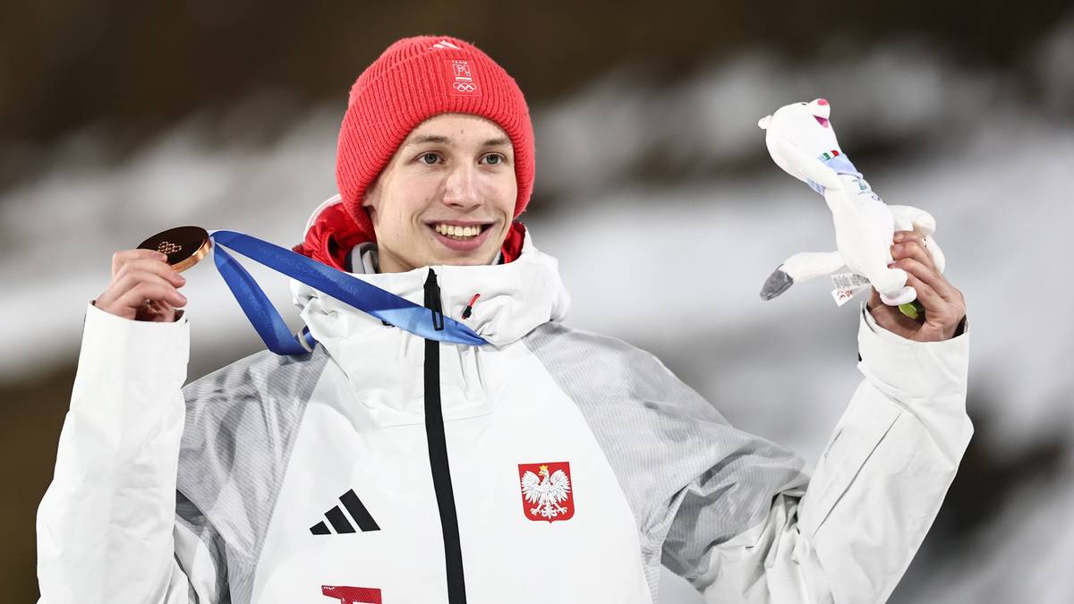Uśmiechnięty skoczek narciarski w czerwonej czapce i kurtce reprezentacji Polski prezentuje brązowy medal i białą maskotkę.