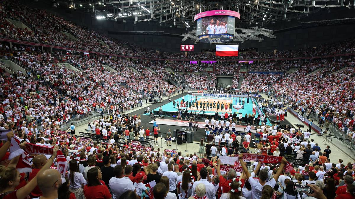 Zdjęcie przedstawia wypełniony po brzegi stadion podczas meczu siatkówki. Na pierwszym planie widać fragment trybun z widzami w biało-czerwonych strojach. Na boisku, otoczonym przez publiczność, znajdują się dwie drużyny siatkarskie, przygotowujące się do ceremonii wręczenia nagród.