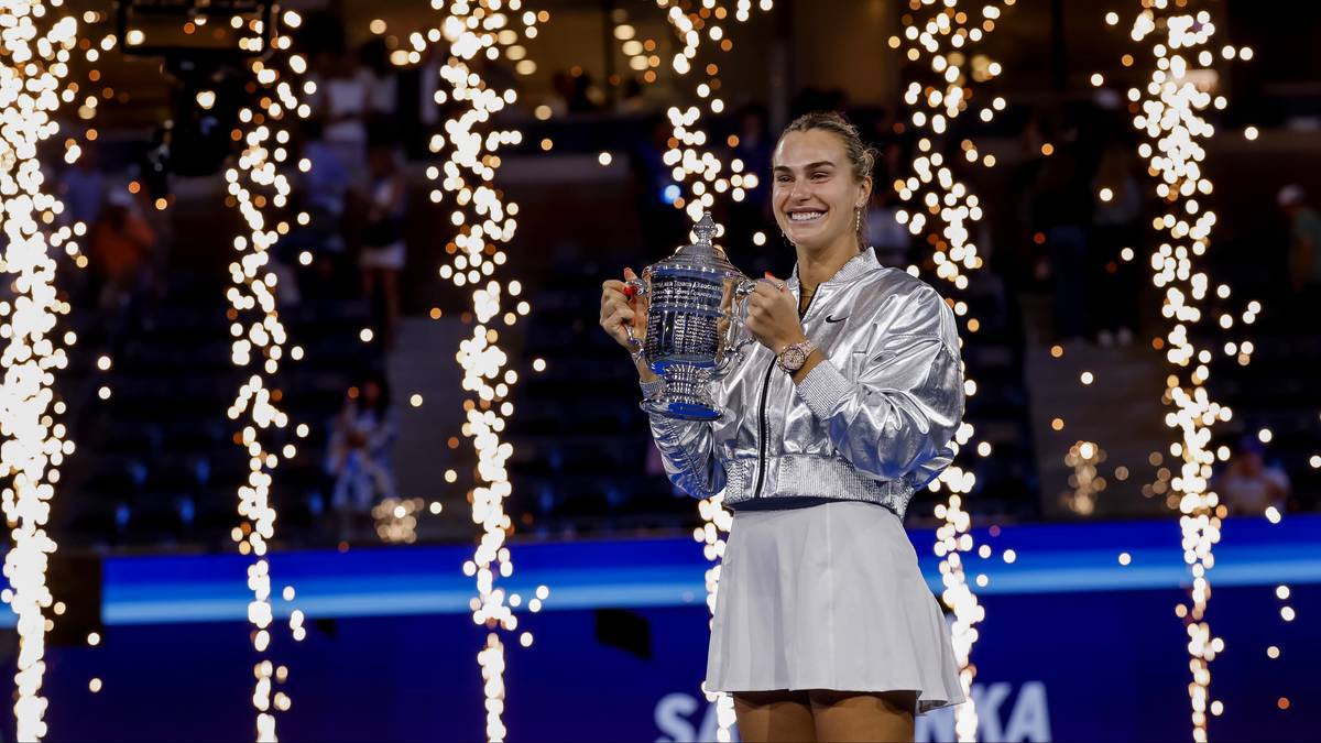 Aryna Sabalenka w srebrnej kurtce i białej spódnicy trzyma trofeum po wygranym finale. Za nią fajerwerki.