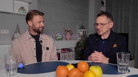 Dwóch mężczyzn siedzi przy stole. Na stole znajdują się owoce i szklanki z napojem. W tle widać półkę z dekoracjami.