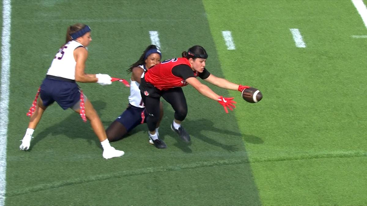 Trzy zawodniczki w trakcie meczu futbolu flagowego. Jedna zawodniczka biegnie z piłką, druga pr&oacute;buje ją zatrzymać, a trzecia jest na ziemi.