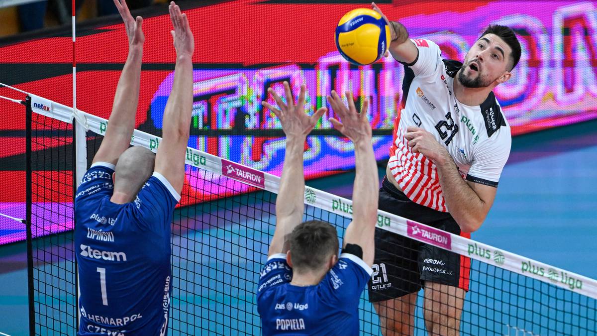 Siatkarz w białej koszulce z czerwonymi pasami wykonuje atak nad siatką, blokowany przez dwóch zawodników w niebieskich strojach.