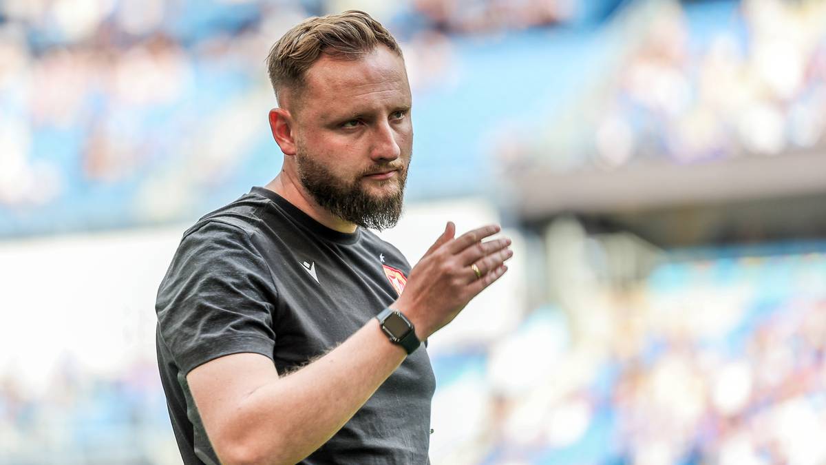 Trener piłkarski w czarnej koszulce z widocznym logo klubu, gestykulujący ręką na tle trybun stadionu.