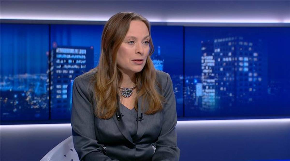 Kobieta w szarej marynarce siedzi i m&oacute;wi, na tle ekranu z nocnym widokiem miasta.