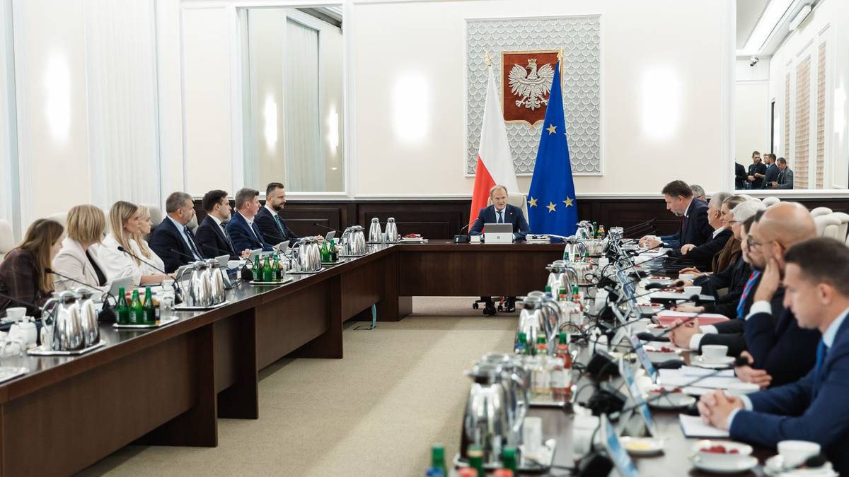 Ludzie siedzący przy długim stole podczas spotkania, z flagami Polski i Unii Europejskiej w tle.