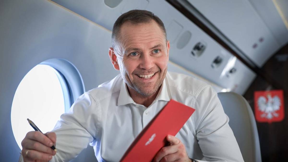 Prezydent Karol Nawrocki siedzi w fotelu w samolocie, trzymając długopis i tablet, z uśmiechem na twarzy.