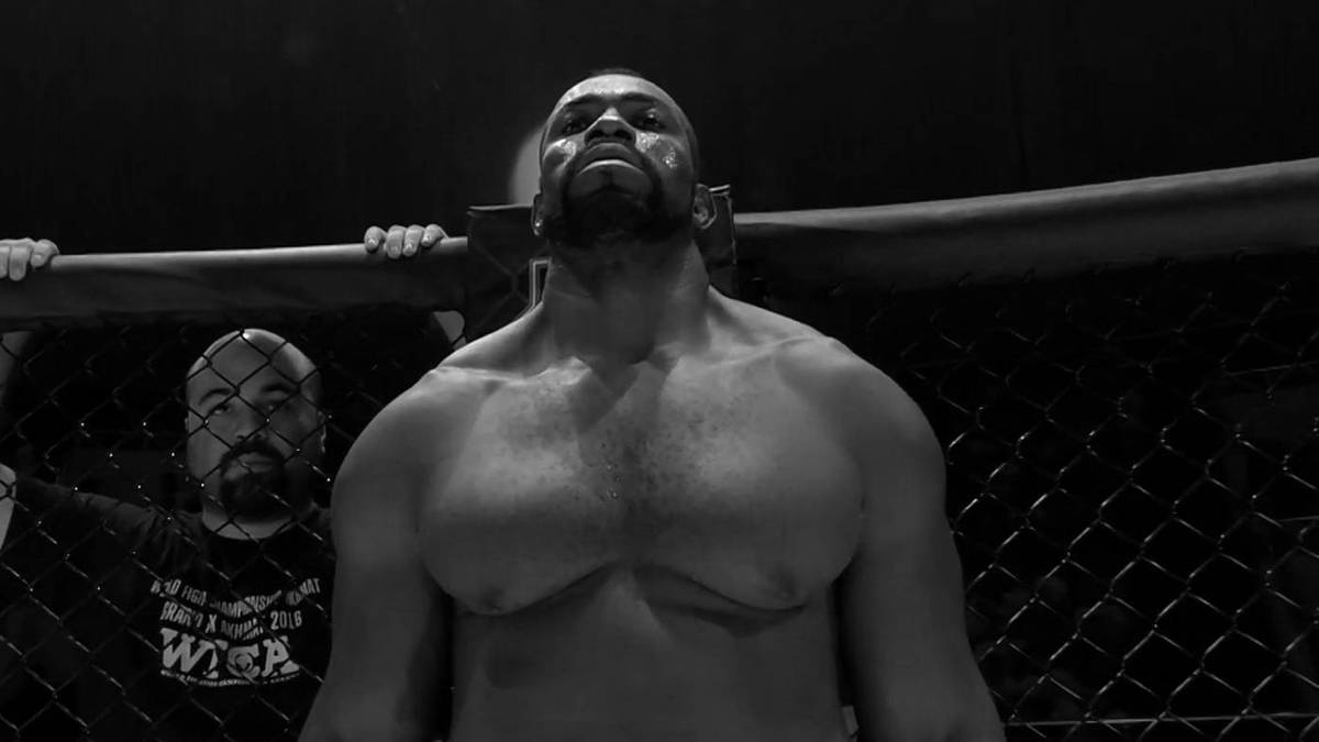 Czarno-białe zdjęcie zawodnika MMA Geronimo Dos Santosa stojącego w klatce, patrzącego przed siebie z podniesioną głową. W tle widoczna jest sylwetka innej osoby.