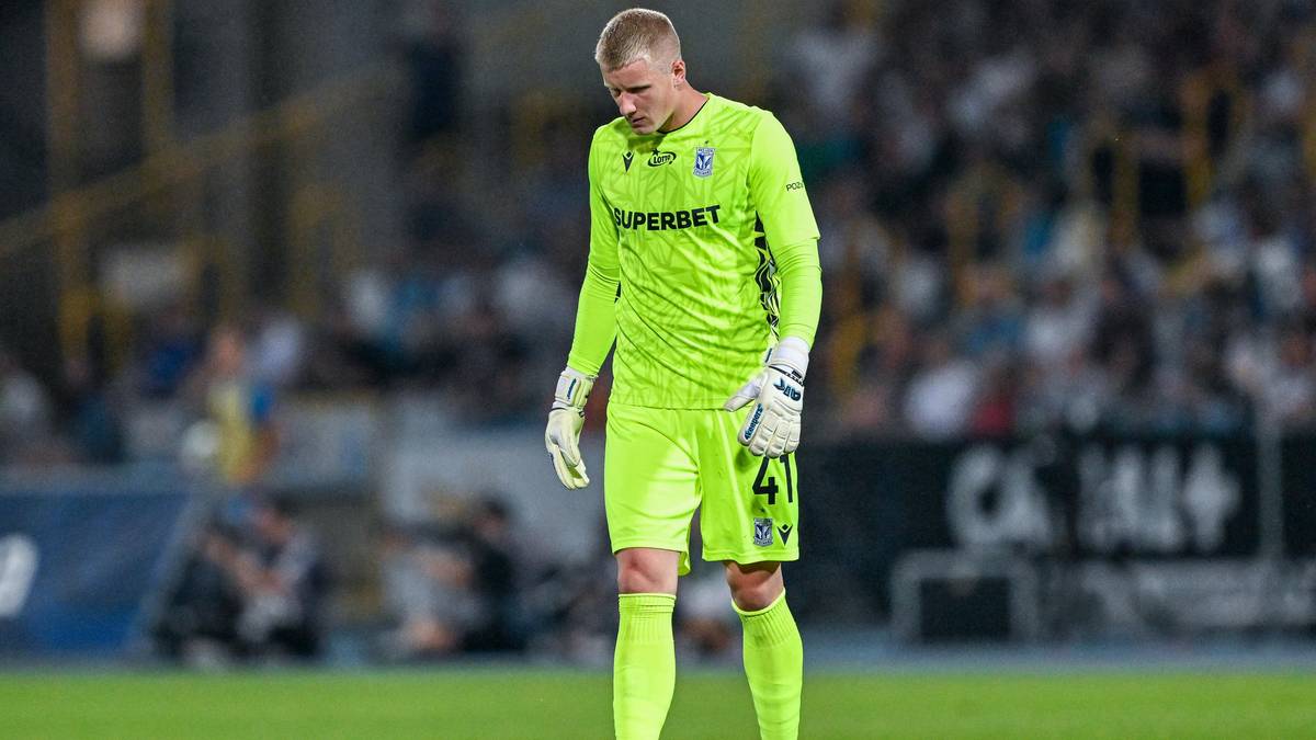 Bramkarz w neonowo-zielonym stroju z numerem 41 na spodenkach stoi na boisku piłkarskim, patrząc w dół.