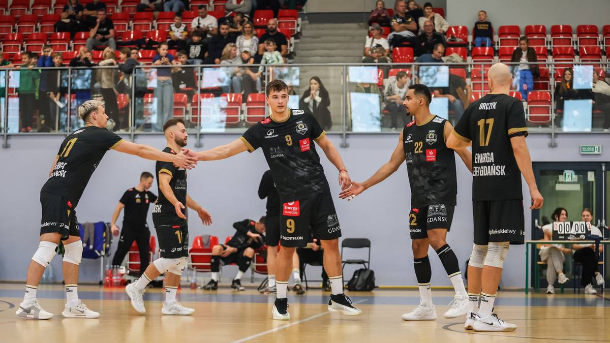 Sześciu siatkarzy w czarnych strojach sportowych z logo Energa Trefl Gdańsk podaje sobie ręce na drewnianym parkiecie hali sportowej. W tle widać czerwone trybuny z widzami.