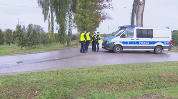 Grupa policjant&oacute;w w kamizelkach odblaskowych stoi obok radiowozu na drodze przy sadzie.