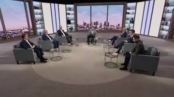 Siedmiu mężczyzn w garniturach siedzi w półokręgu na fotelach w studiu telewizyjnym. Za nimi znajdują się panoramiczne okna z widokiem na miasto.