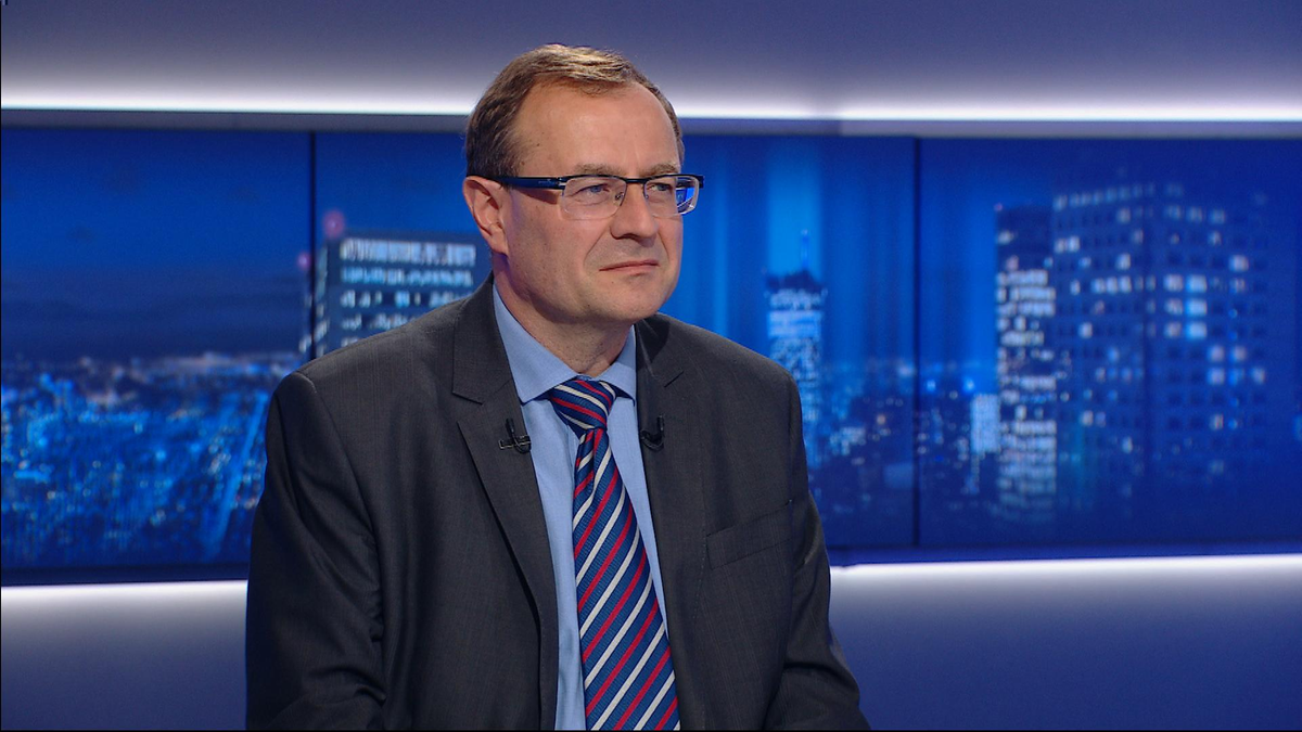 Mężczyzna w garniturze i krawacie siedzi przed ekranami z widokiem nocnego miasta.