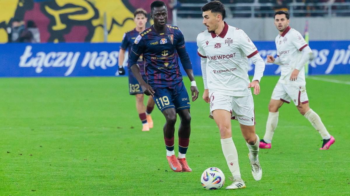 Piłkarz w białym stroju prowadzi piłkę, a obok niego dwaj przeciwnicy w granatowych strojach próbują odebrać mu futbolówkę.