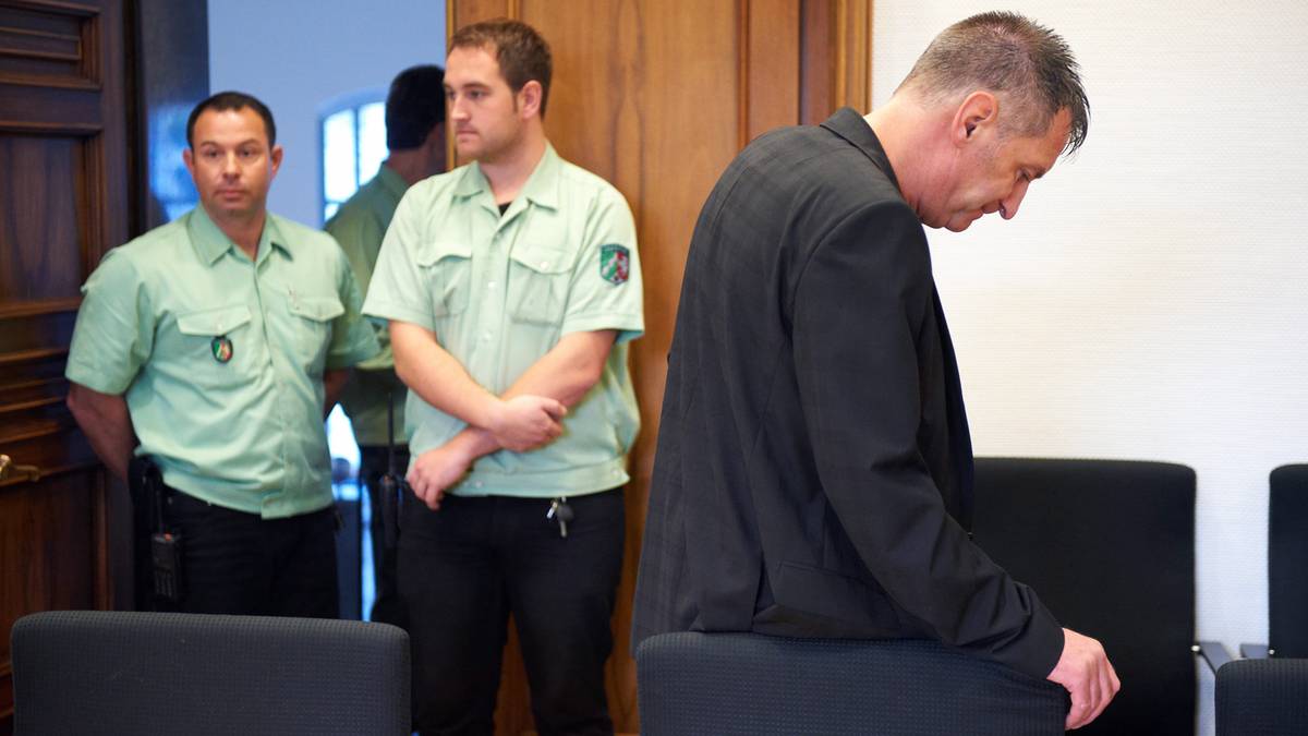 Mężczyzna w czarnej marynarce spuszcza głowę, stojąc w sądzie przed dwoma strażnikami w jasnych koszulach.