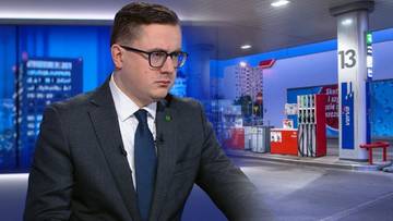 Mężczyzna w garniturze i okularach przed ekranem z grafiką stacji paliw.