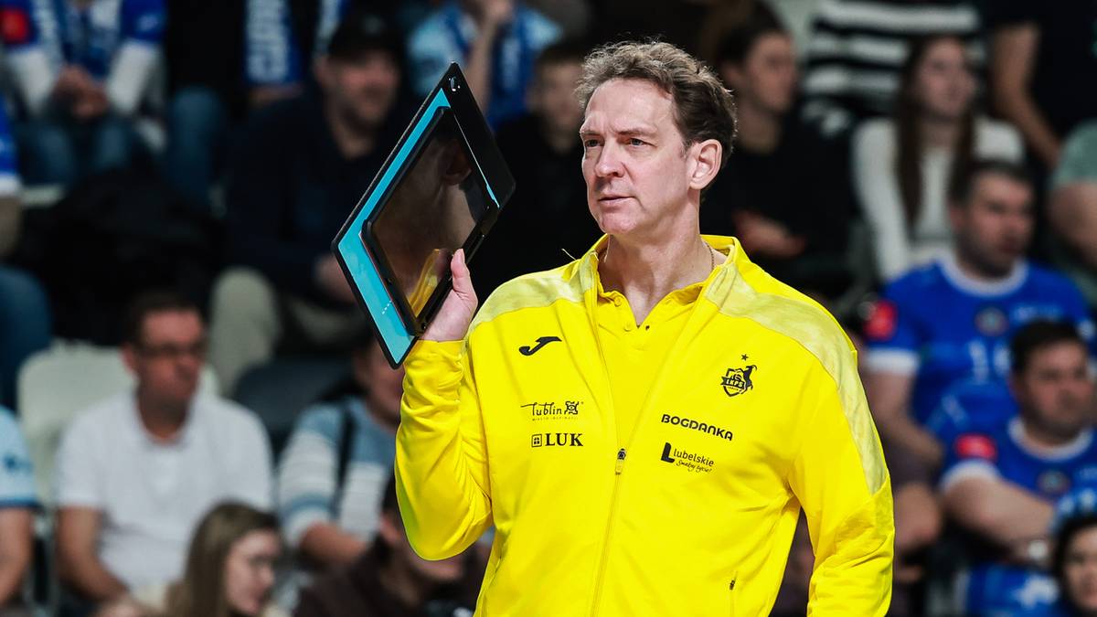 Trener Stephane Antiga w ż&oacute;łtej kurtce treningowej z tabletem w ręku, stojący na tle trybun wypełnionych kibicami na hali sportowej.