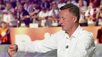 Tomasz Hajto w białej koszuli, gestykulujący prawą ręką podczas wypowiedzi. W tle widoczne są trybuny stadionu wypełnione kibicami.