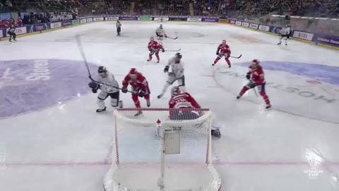 Zawodnicy drużyn IFK Helsinki i Sparta Praga grają w hokeja. Akcja toczy się pod bramką, gdzie jeden z zawodnik&oacute;w w czerwonym stroju strzela, a bramkarz w białym stroju pr&oacute;buje obronić. Inni zawodnicy w czerwonych i białych strojach znajdują się w pobliżu.