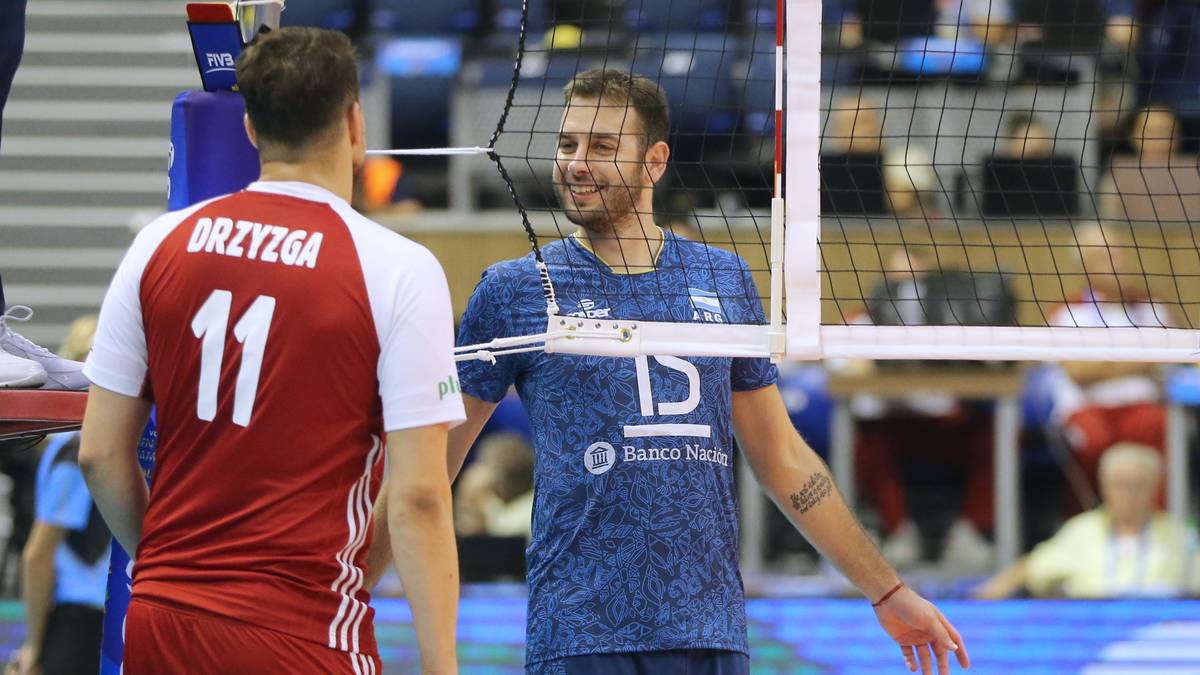 Dwóch siatkarzy na boisku, jeden w czerwonej koszulce z numerem 11 i napisem "Drzyzga", drugi w niebieskiej koszulce z logo klubu "Banco Nacionale". Siatkarze stoją przy siatce i rozmawiają.