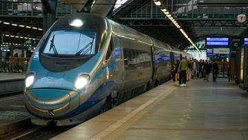 Nowoczesny, niebiesko-srebrny pociąg ekspresowy zatrzymany przy peronie na zadaszonej stacji kolejowej, z widocznym tłumem podr&oacute;żnych wsiadających do wagon&oacute;w.