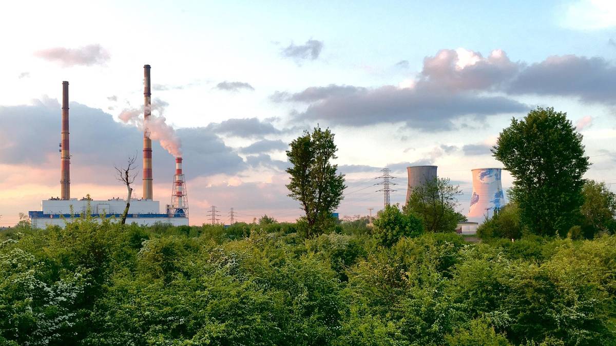 Widok elektrowni z dymiącymi kominami na tle zachodzącego słońca, otoczonej zielenią.