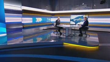 Dw&oacute;ch mężczyzn w studiu telewizyjnym, jeden rozmawia z drugim przy okrągłym stole. Na ekranie w tle widnieje napis "graffiti".