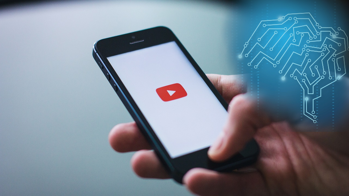 Dłoń trzymająca smartfon z otwartą aplikacją YouTube, obok znajduje się grafika przedstawiająca m&oacute;zg z obwodami elektronicznymi, symbolizująca sztuczną inteligencję.