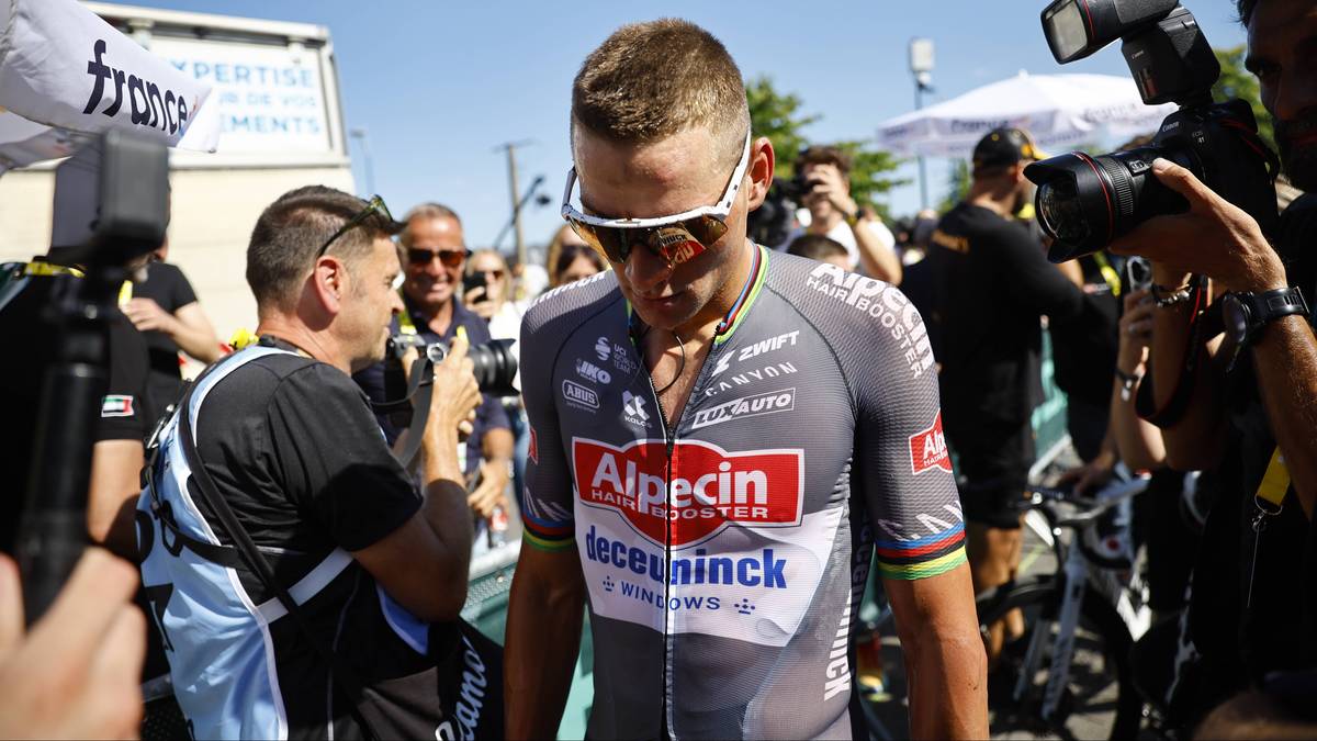 Kolarz w szarym stroju z logo Alpecin-Deceuninck i okularach sportowych na nosie, otoczony przez ludzi i fotoreporter&oacute;w.