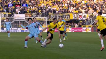 Dw&oacute;ch piłkarzy walczy o piłkę na boisku podczas meczu NAC Breda - Feyenoord Rotterdam. W tle widoczni inni zawodnicy, bramka oraz trybuny z kibicami.