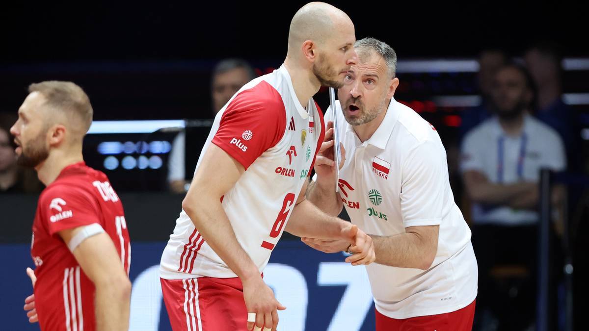 Siatkarz reprezentacji Polski w białej koszulce z czerwonymi pasami rozmawia z trenerem w białej koszulce z zielonymi elementami.