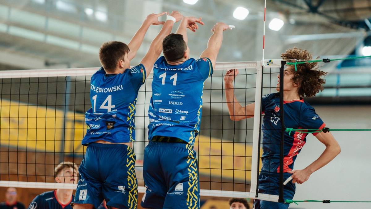 Siatkarze w niebieskich strojach walczą przy siatce, pr&oacute;bując zablokować lub wykonać atak podczas meczu.