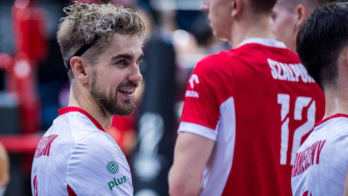 Młody siatkarz o jasnych, kręconych włosach i brodzie, w białej koszulce z zielonym logo, uśmiechnięty, patrzy w bok. Za nim stoją inni siatkarze w czerwonych koszulkach.