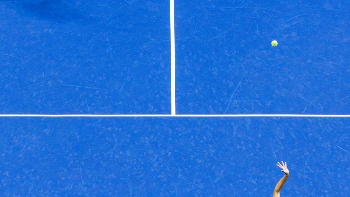 Kobieta w białej bluzce i kr&oacute;tkiej sp&oacute;dniczce rzuca do serwisu piłeczkę tenisową na niebieskim korcie.